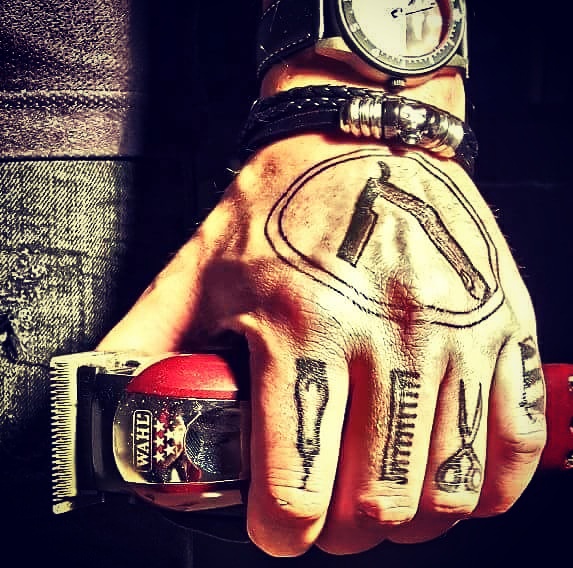 Een hand met getatoeëerde kappersgereedschappen op de vingers houdt een tondeuse vast